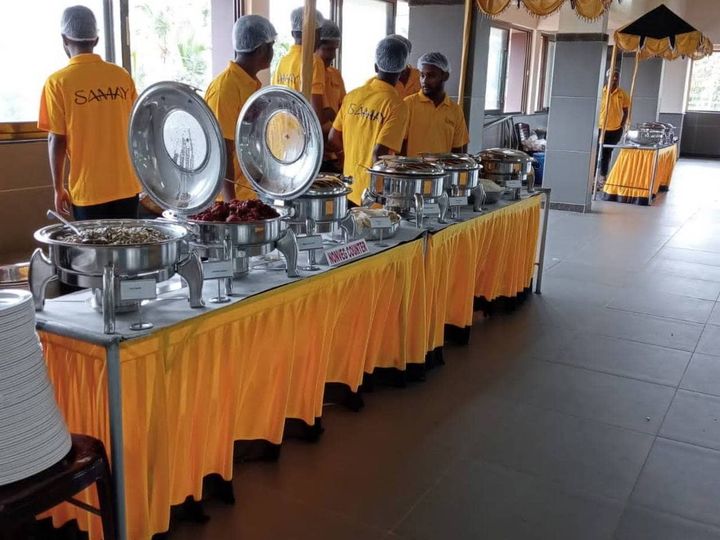 Catering staff in yellow Samay t-shirts and hairnets at a long buffet table with steel chafing dishes