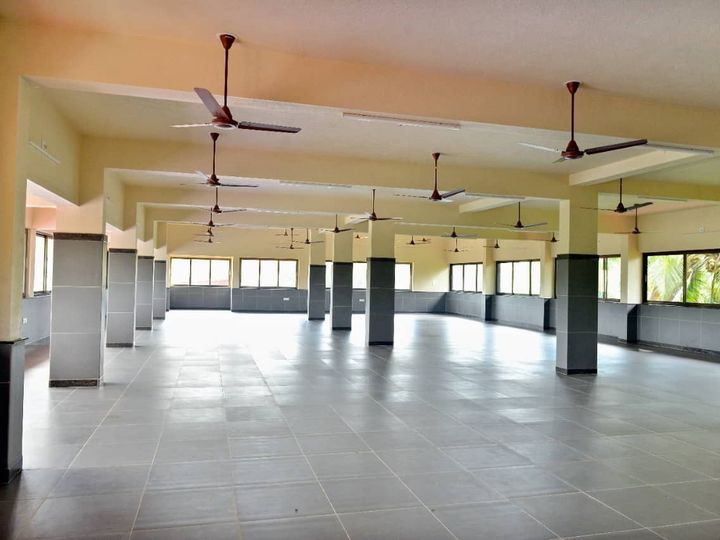 Wide hall with a grey tiled floor, square pillars, ceiling fans and windows on every side