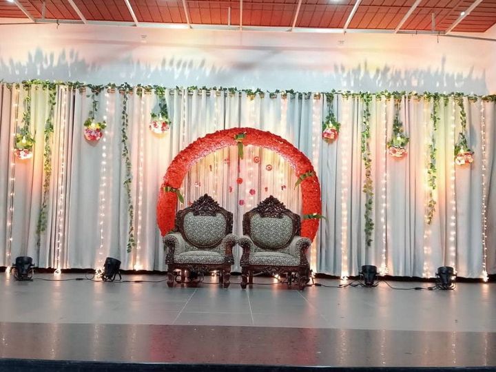 Stage with two carved chairs in front of a red floral ring, white curtains, hanging flowers and fairy lights